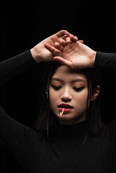 portrait of pretty asian model with burning match in mouth posing isolated on black - 写真・画像