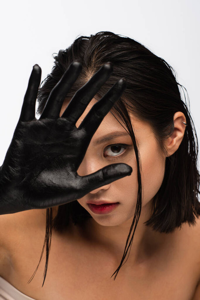 young asian woman with bare shoulders covering face with hand in black paint isolated on white - Foto, Imagen