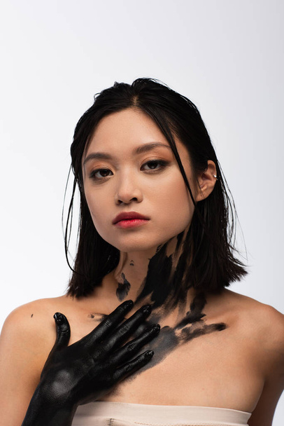 young asian woman with bare shoulders and hand in black paint touching body while looking at camera isolated on white - Photo, image
