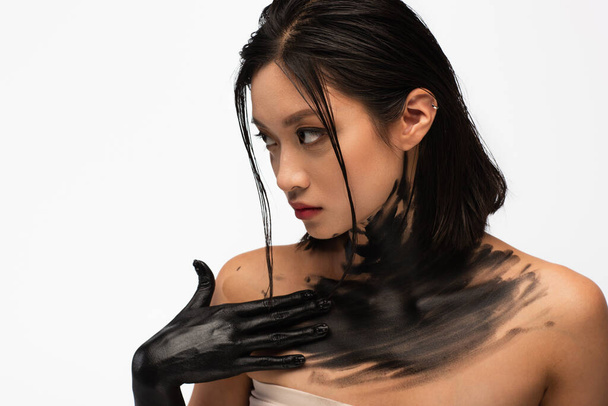 young asian woman with naked shoulders and hand in black paint touching body isolated on white - Photo, image