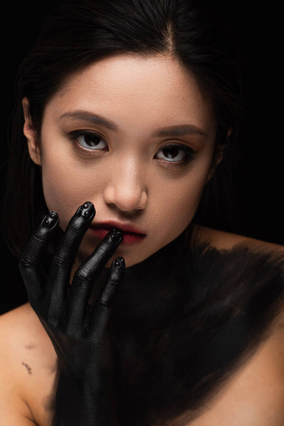 young asian woman with bare shoulders and hand in paint looking at camera while touching lip isolated on black - Φωτογραφία, εικόνα
