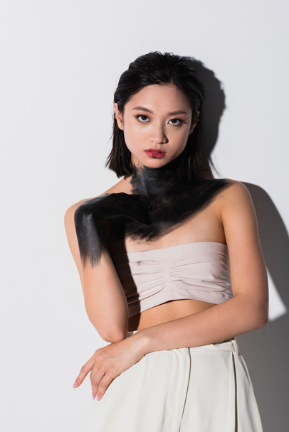 young asian woman with bare shoulders and hand in black paint looking at camera on white - Foto, Bild