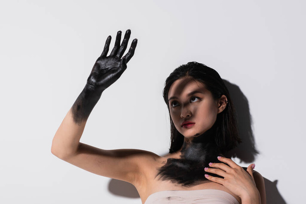 young asian woman holding hand in black paint above face while covering from light on white - Foto, afbeelding