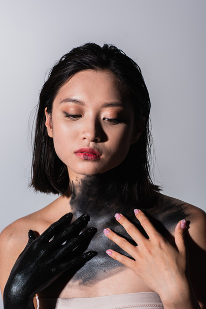 молодая азиатская женщина с голыми плечами и телом в черной краске изолированы на сером - Фото, изображение