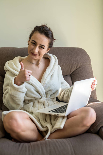 mooi jong meisje in een sneeuwwitte badjas met een laptop op haar knieën maakt online winkelen. Online winkelen concept. - Foto, afbeelding