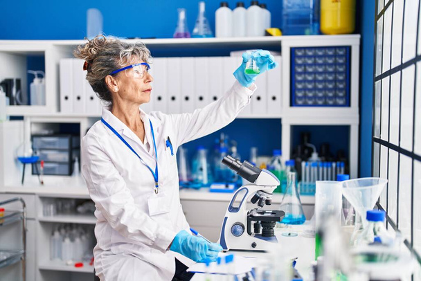 Middle age woman scientist holding test tube write on clipboard at laboratory - Photo, Image