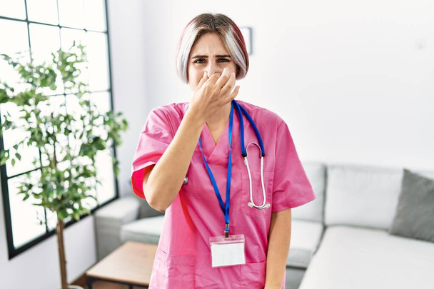 Young beautiful woman wearing doctor uniform and stethoscope smelling something stinky and disgusting, intolerable smell, holding breath with fingers on nose. bad smell  - Foto, Bild