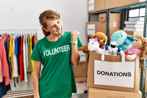 Beautiful caucasian woman wearing volunteer t shirt at donations stand very happy and excited doing winner gesture with arms raised, smiling and screaming for success. celebration concept.  - Photo, Image