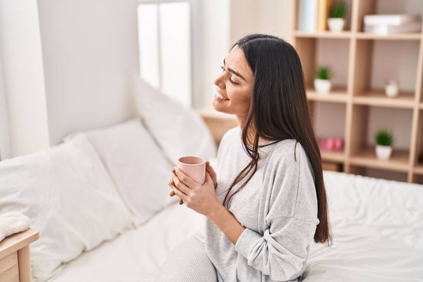 Young hispanic woman drinking cup of coffee sitting on bed at bedroom - Foto, afbeelding