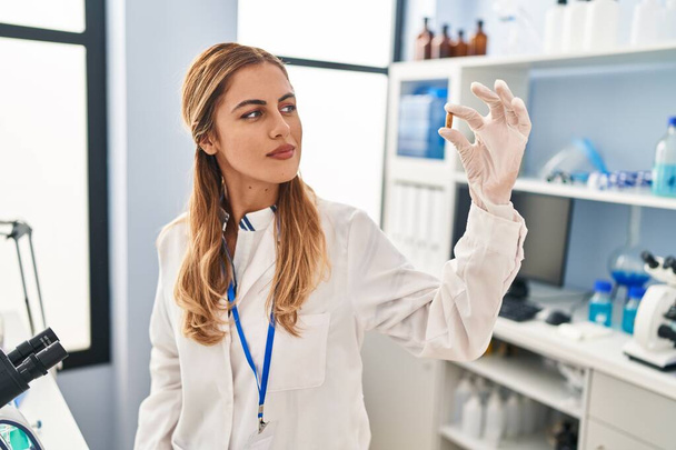 Young blonde woman wearing scientist uniform holding pill at laboratory - Fotó, kép