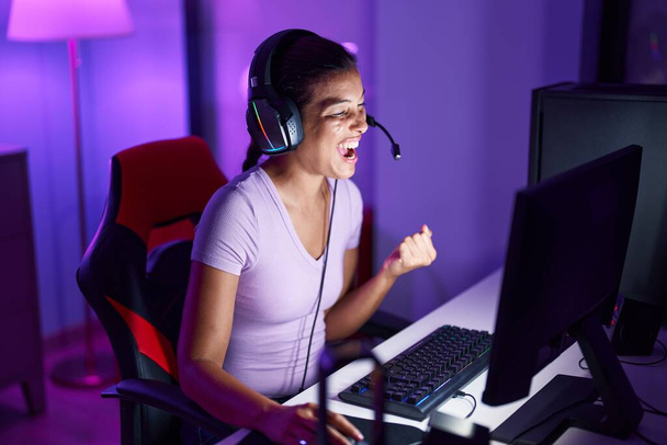 Νεαρή όμορφη ισπανόφωνη γυναίκα streamer παίζει βιντεοπαιχνίδι με την έκφραση νικητής στην αίθουσα τυχερών παιχνιδιών - Φωτογραφία, εικόνα