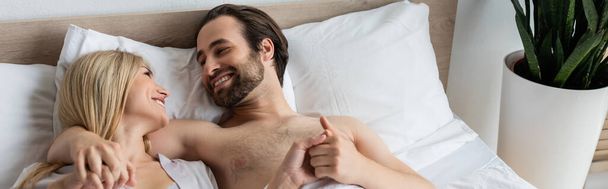 χαμογελαστό ζευγάρι κρατώντας τα χέρια και κοιτάζοντας ο ένας τον άλλον, ενώ βρίσκονται στο κρεβάτι, πανό - Φωτογραφία, εικόνα