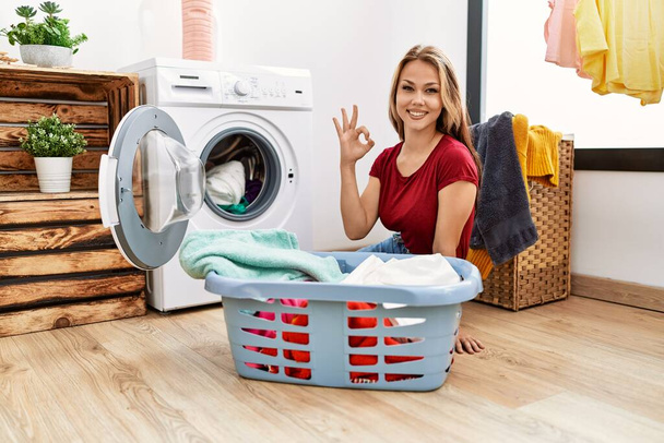 Молода біла жінка вкладає брудну білизну в пральну машину, роблячи знак ок з пальцями, посміхаючись дружній жест відмінний символ
  - Фото, зображення