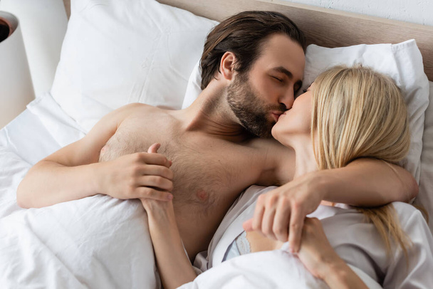 high angle view of blonde woman and brunette man holding hands and kissing in bed - Foto, imagen