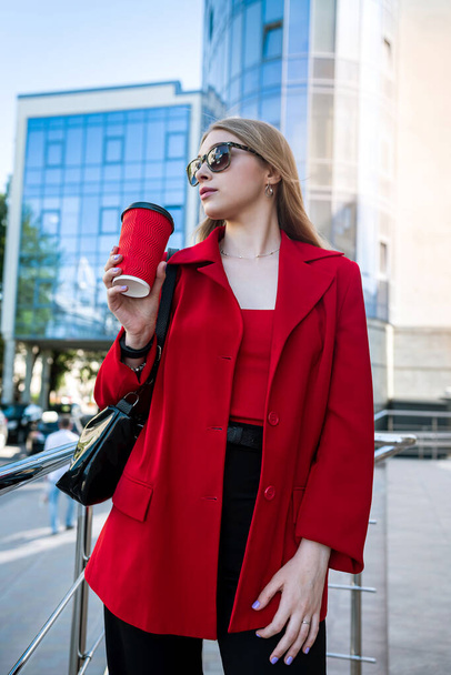 A business woman with a cup of coffee is talking on her smartphone while standing in the middle of the city center. The concept of a woman who earns money - Zdjęcie, obraz