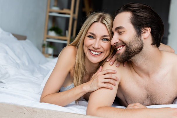 Ευτυχισμένη ξανθιά γυναίκα χαμογελά στην κάμερα και αγκαλιάζει γυμνό άντρα στο κρεβάτι - Φωτογραφία, εικόνα