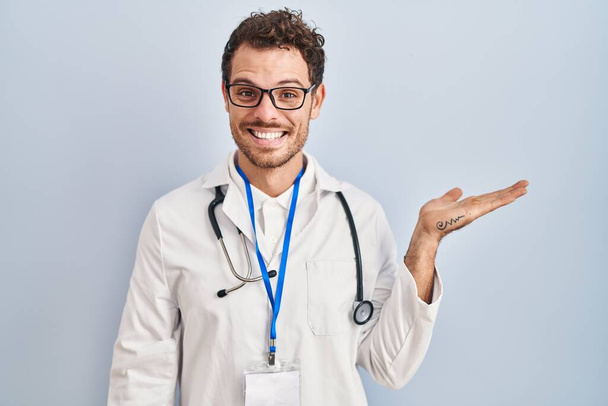 Junger hispanischer Mann in Arztuniform und Stethoskop lächelt fröhlich und zeigt mit der Handfläche in die Kamera.  - Foto, Bild