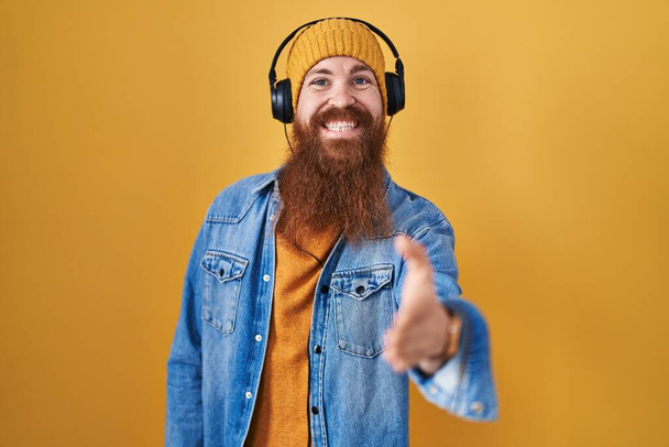 Hombre caucásico con barba larga escuchando música usando auriculares sonrientes amistosos ofreciendo apretón de manos como saludo y bienvenida. negocios exitosos.  - Foto, imagen