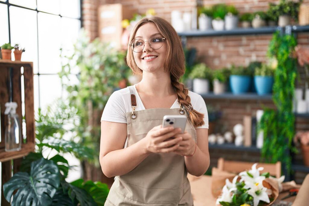 Joven florista rubia sonriendo confiada usando teléfono inteligente en floristería - Foto, imagen