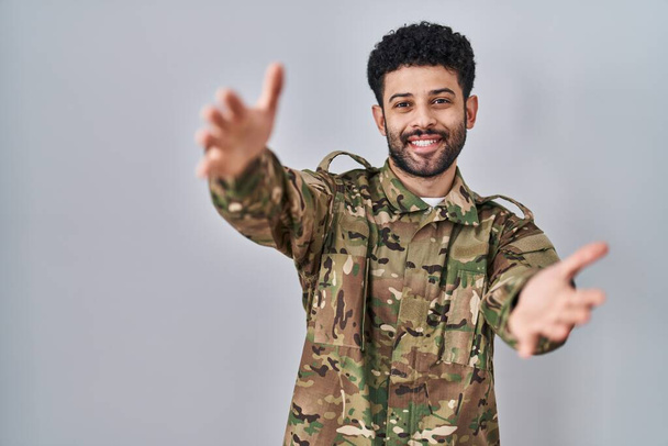 Homme arabe portant un uniforme de camouflage de l'armée regardant la caméra souriant à bras ouverts pour étreindre. expression joyeuse embrassant le bonheur.  - Photo, image