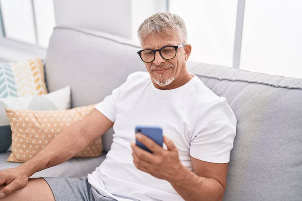 Middle age grey-haired man using smartphone sitting on sofa at home - Foto, Imagem