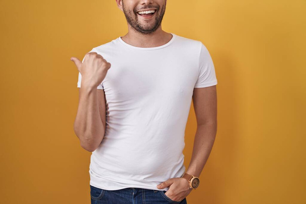 Hispanic man wearing white t shirt over yellow background pointing thumb up to the side smiling happy with open mouth  - Foto, Imagen
