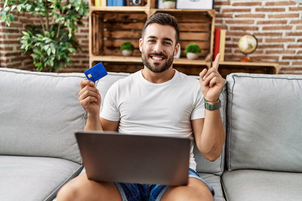 Jóképű spanyol férfi csinál online vásárlás gazdaság hitelkártya mosolyogva boldog mutató kéz és ujj az oldalon  - Fotó, kép