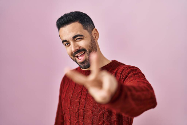 Νεαρός Ισπανός με γενειάδα που φοράει ζακέτα πάνω από ροζ φόντο χαμογελώντας κοιτάζοντας την κάμερα που δείχνει τα δάχτυλα να κάνουν το σήμα της νίκης. Νούμερο δύο..  - Φωτογραφία, εικόνα