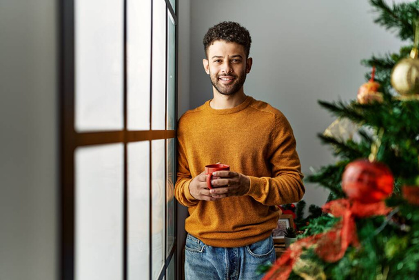 Young arab man drinking coffee standing by christmas tree at home. - Photo, Image