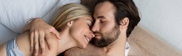 top view of brunette man hugging smiling woman while lying on bed with closed eyes, banner - Foto, imagen