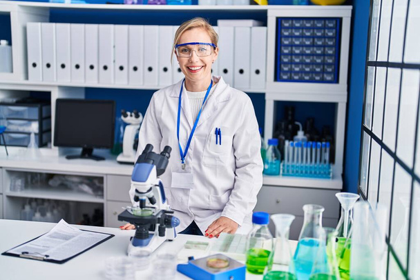 Jonge blonde vrouw wetenschapper glimlachen zelfverzekerd met behulp van microscoop in laboratorium - Foto, afbeelding