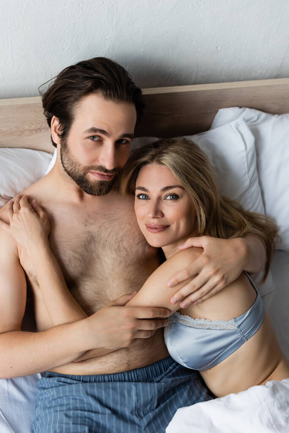 top view of smiling lovers embracing and looking at camera on bed - Photo, Image