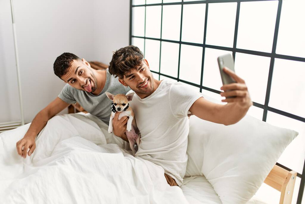 Δύο Ισπανοί άνδρες ζευγάρι κάνουν selfie από το smartphone κάθεται στο κρεβάτι με chihuahua στο υπνοδωμάτιο - Φωτογραφία, εικόνα