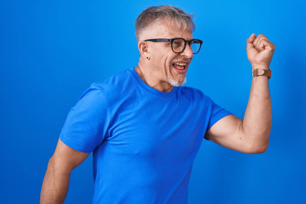 Homem hispânico com cabelos grisalhos em pé sobre fundo azul dançando feliz e alegre, sorrindo em movimento casual e confiante ouvir música  - Foto, Imagem
