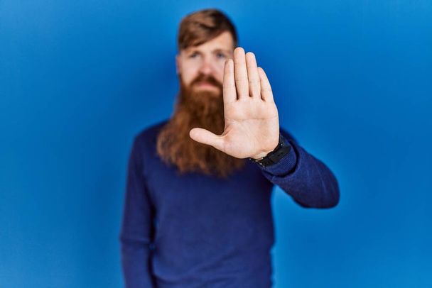 Roodharige man met lange baard draagt casual blauwe trui over blauwe achtergrond doen stoppen met zingen met palm van de hand. waarschuwingsuitdrukking met negatief en ernstig gebaar op het gezicht.  - Foto, afbeelding