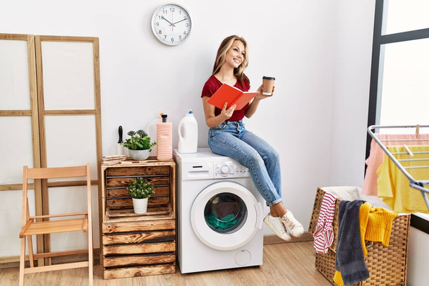 Young caucasian girl drinking coffee and reading book waiting for laundry sitting on whasing machine at home. - Foto, Imagem