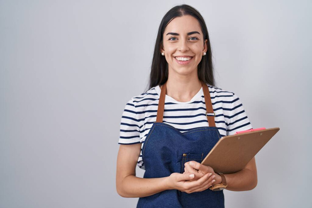 Young brunette woman wearing professional waitress apron and clipboard looking positive and happy standing and smiling with a confident smile showing teeth  - Φωτογραφία, εικόνα