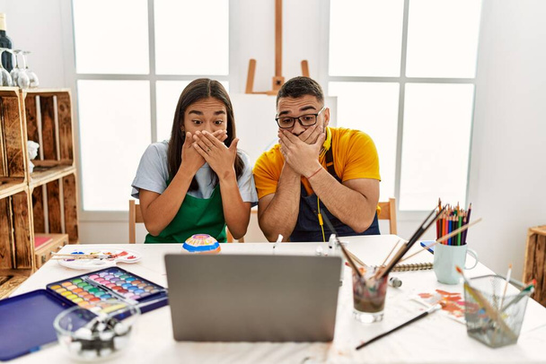 Young hispanic couple using laptop sitting on the table at art studio shocked covering mouth with hands for mistake. secret concept.  - Fotografie, Obrázek