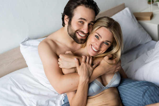 χαρούμενο και σέξι ζευγάρι αγκαλιάζει και χαμογελά στην κάμερα στο κρεβάτι - Φωτογραφία, εικόνα