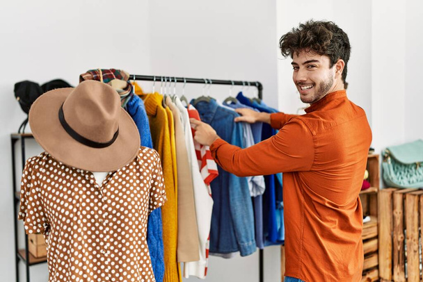 Joven cliente hispano sonriendo feliz eligiendo ropa en la tienda de ropa. - Foto, imagen