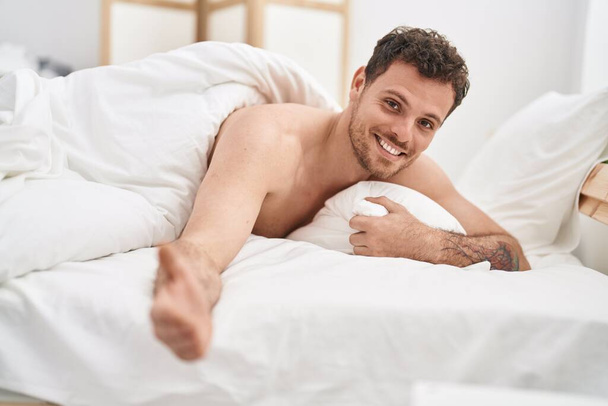 Νεαρός Ισπανός άντρας χαμογελάει με αυτοπεποίθηση ξαπλωμένος στο κρεβάτι στην κρεβατοκάμαρα - Φωτογραφία, εικόνα