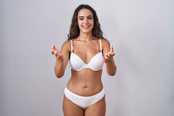 jonge hispanic vrouw het dragen van witte lingerie glimlachend vrolijk aanbieden van handen geven van hulp en acceptatie.  - Foto, afbeelding