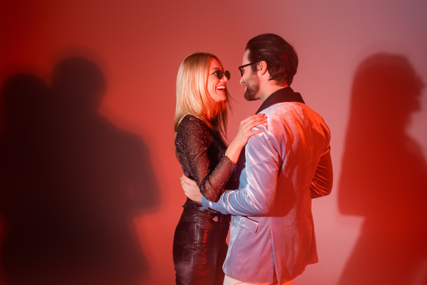 Cheerful woman in sunglasses hugging stylish boyfriend on red background - Photo, image