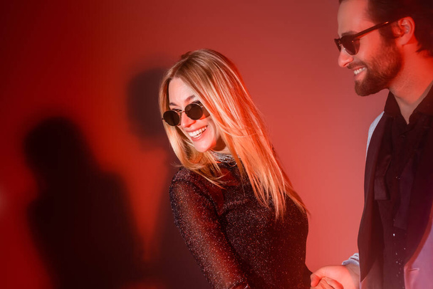 Улыбающийся мужчина в солнечных очках смотрит на веселую девушку во время вечеринки на красном фоне - Фото, изображение
