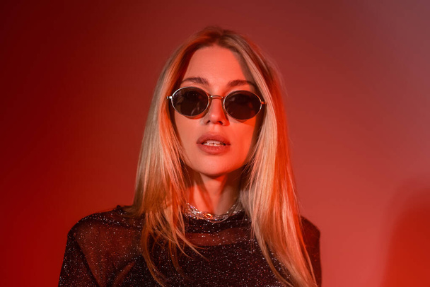 Stylish blonde woman in sunglasses standing on red background  - Foto, Imagen