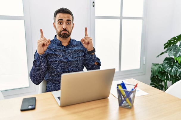 Joven hispano con barba trabajando en la oficina con laptop apuntando hacia arriba mirando triste y molesto, indicando dirección con los dedos, infeliz y deprimido.  - Foto, Imagen