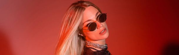 Trendy woman in sunglasses looking at camera on red background, banner  - Fotografie, Obrázek