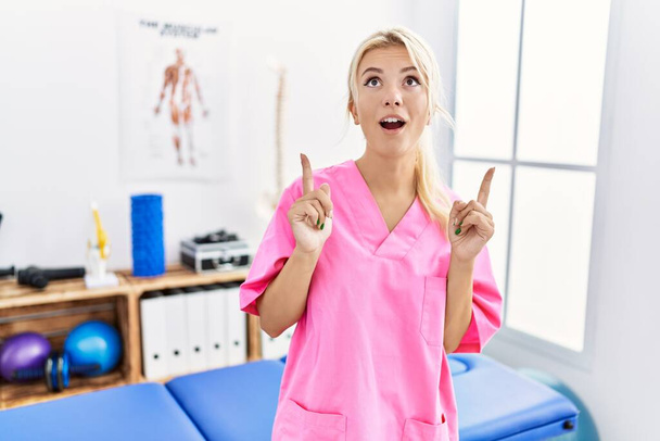 Young caucasian woman working at pain recovery clinic amazed and surprised looking up and pointing with fingers and raised arms.  - Photo, Image