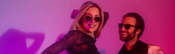 Smiling woman in sunglasses dancing with boyfriend during party on purple background, banner   - Fotó, kép