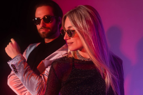 Stylish woman in sunglasses standing near blurred boyfriend during party on purple background  - Foto, imagen